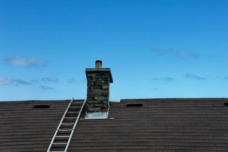 A ladder on a tiled roof next to a chimney, under a blue sky
