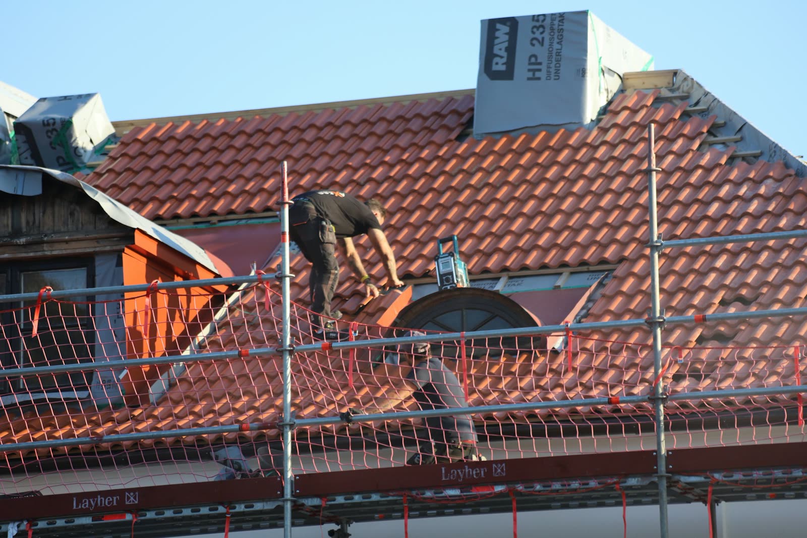 A roofer laying red clay roof tiles behind scaffolding on a sunny day